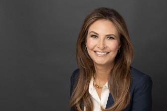 Studio portrait shows a business professional wearing a dark blazer over a light blouse, seated against a plain gray backdrop with soft, even lighting