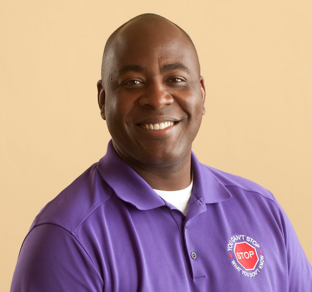 Person wearing purple polo shirt with white undershirt; shirt features red stop sign logo and text reading ‘You Can’t Stop What You Don’t Know