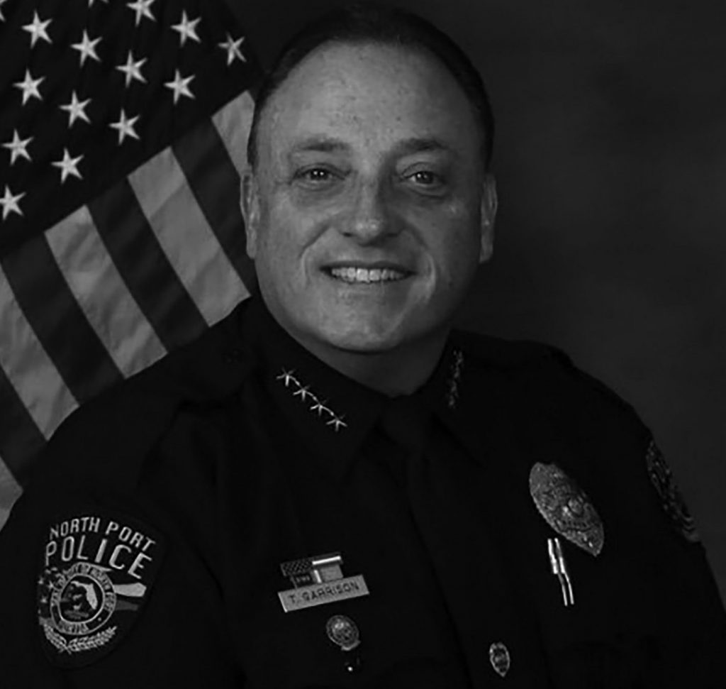 A uniformed police officer is seated in front of an American flag. The uniform includes a badge, name tag, and several pins on the chest. The shoulder patch reads "North Port Police." The officer wears a tie and has four stars on the collar, indicating rank.