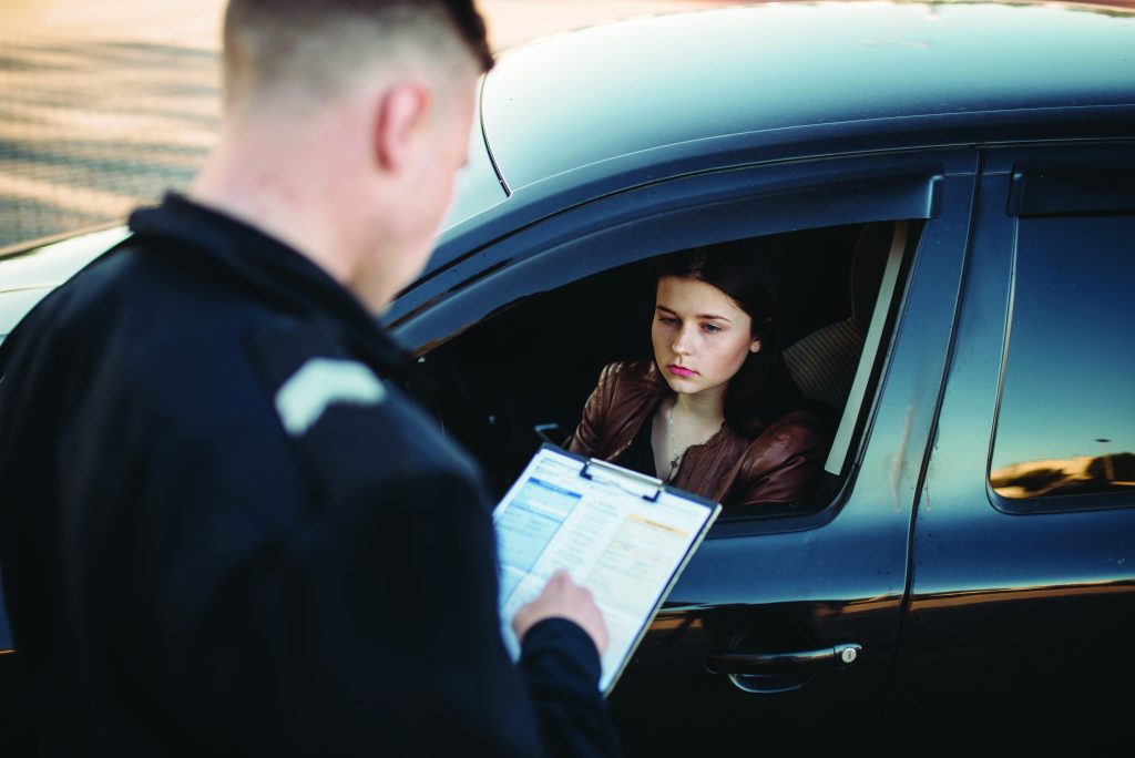 Officer stands beside a stopped vehicle holding a clipboard with forms while the driver sits in the car window, during a daylight roadside check
