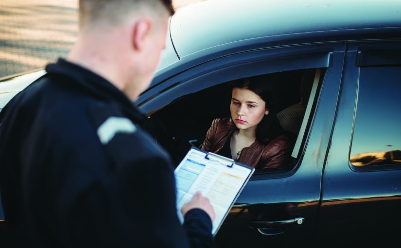 Officer stands beside a stopped vehicle holding a clipboard with forms while the driver sits in the car window, during a daylight roadside check