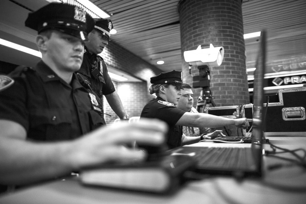 Uniformed police officers work at folding tables with laptops, radios, and video equipment inside a brick transit station, using tripods, cables, and control cases