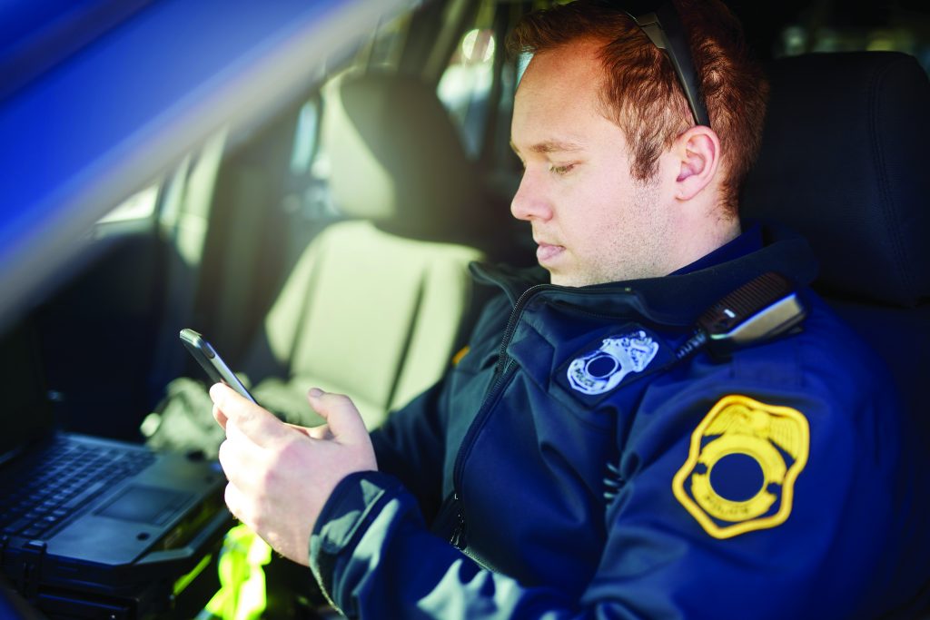 a police officer seated inside a vehicle, wearing a dark blue uniform with a badge on the left shoulder. The officer's face is obscured by a pixelated square. The officer is holding a smartphone in the right hand and appears to be looking at it. There is an open laptop on the center console of the vehicle, positioned to the left of the officer. Sunlight is streaming through the window, illuminating part of the officer's face and uniform.