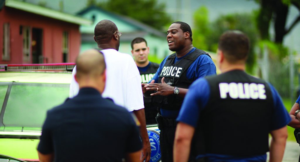 Police officers wearing body armor marked ‘POLICE’ speak with an individual near a patrol vehicle on a residential street lined with houses, trees, and parked cars