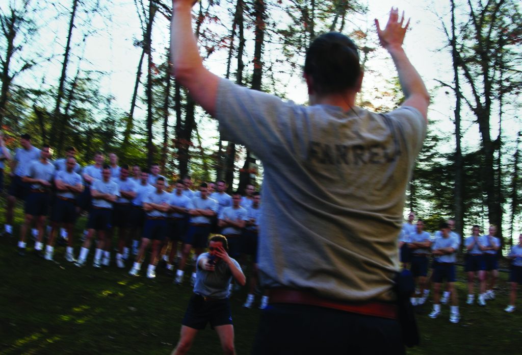 Outdoor training scenario shows raised hands in foreground while a trainee aims a handgun ahead, with group in uniform shirts observing among tall trees and grass