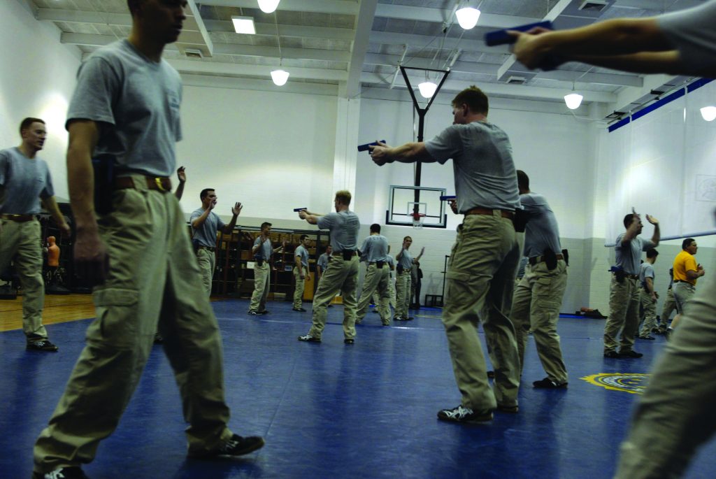 Indoor training scenario in gym shows multiple trainees aiming handguns while participants raise hands, with mats, basketball hoop, lockers, and overhead lights