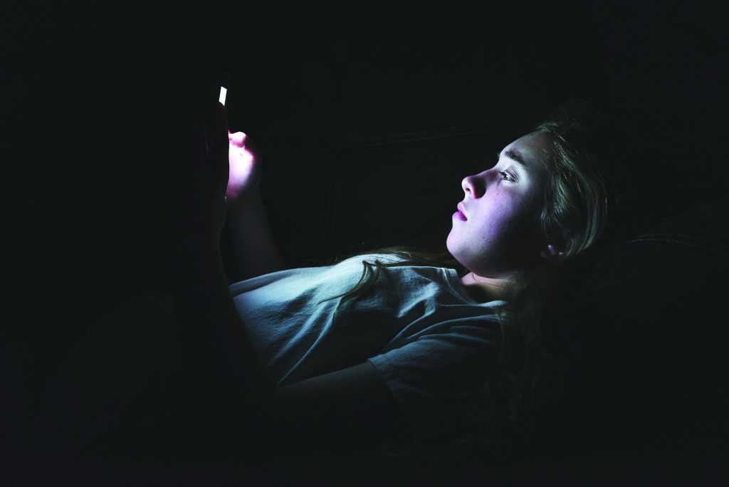 Person in dark room lit by smartphone glow; face obscured, wearing light shirt; scene suggests digital activity or surveillance in low light