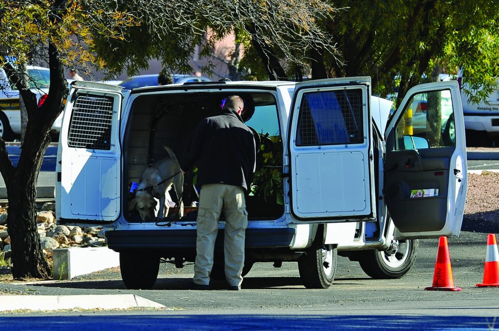 Officer stands at open back of white van with caged doors, interacting with dog inside; side door open, orange traffic cones on asphalt nearby