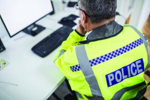 Officer in high-visibility jacket labeled 'POLICE' sits at desk using computer with keyboard and mouse; focused on screen in indoor workspace setting