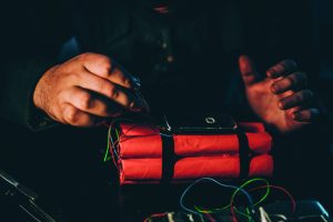 Hands assembling explosive device with red dynamite sticks, black tape, multicolored wires, and mobile phone used as trigger; close-up view