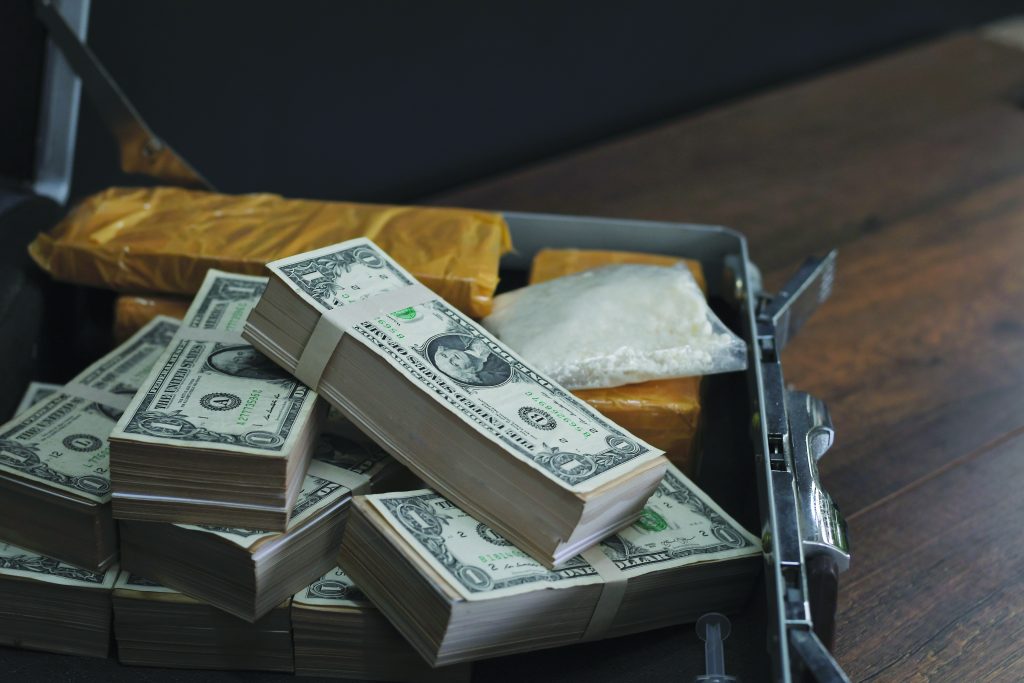 Open briefcase on wooden table with stacks of U.S. bills, yellow-wrapped packages, and clear plastic bag containing white powder; evidence setup