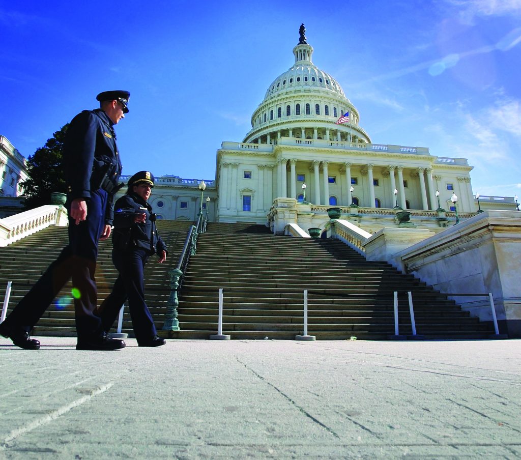 Uniformed Capitol police walk across wide stone steps in front of the U.S. Capitol building, with dome, columns, flag, and clear blue sky in sunlight