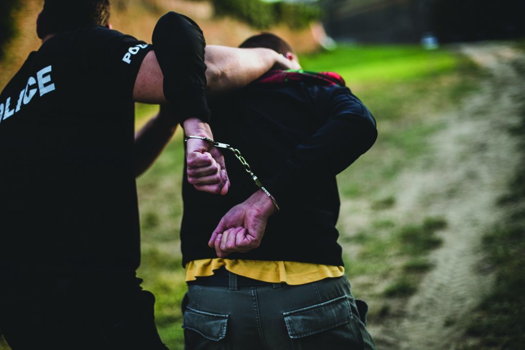 a police officer arresting a person outdoors. The officer, wearing a black shirt with the word "POLICE" in white letters on the back, is holding the person's arm and placing handcuffs on their wrists behind their back. The person being arrested is wearing a black jacket over a yellow shirt and has short hair. The scene takes place on a dirt path with grass and trees in the background. Both individuals' faces are not fully visible as they are turned away from the camera.