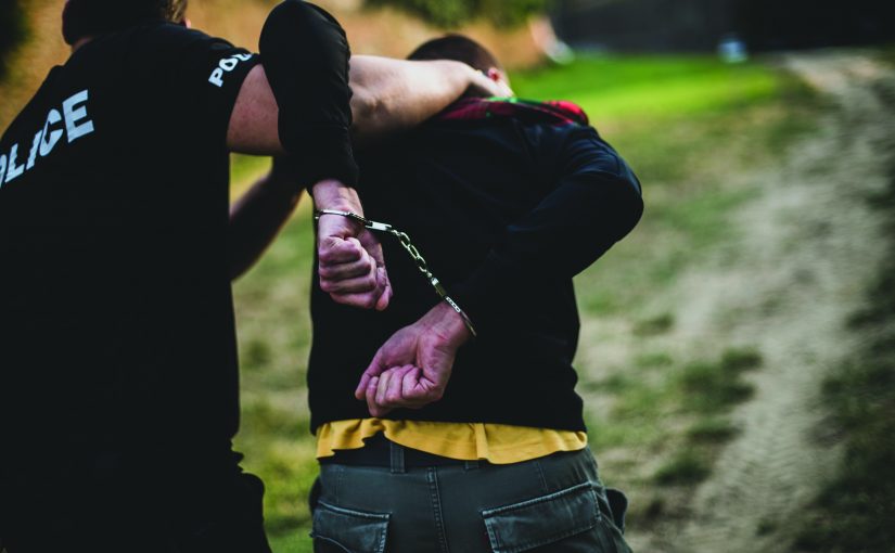 a police officer arresting a person outdoors. The officer, wearing a black shirt with the word "POLICE" in white letters on the back, is holding the person's arm and placing handcuffs on their wrists behind their back. The person being arrested is wearing a black jacket over a yellow shirt and has short hair. The scene takes place on a dirt path with grass and trees in the background. Both individuals' faces are not fully visible as they are turned away from the camera.