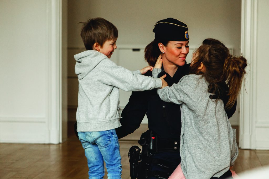 A uniformed police officer kneels inside a residential room while two young children reach toward the officer’s shoulders, with hardwood floors, white walls, and doorways visible