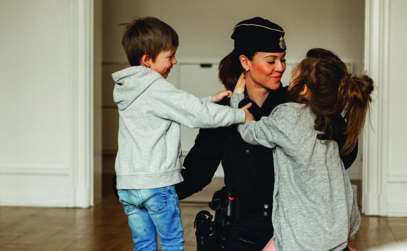 A uniformed police officer kneels inside a residential room while two young children reach toward the officer’s shoulders, with hardwood floors, white walls, and doorways visible