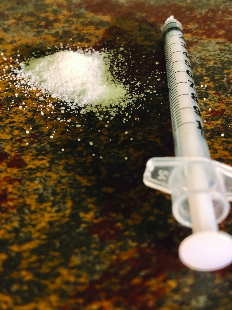 Close-up of white crystalline powder on textured surface beside clear plastic syringe with measurement markings, suggesting narcotics evidence