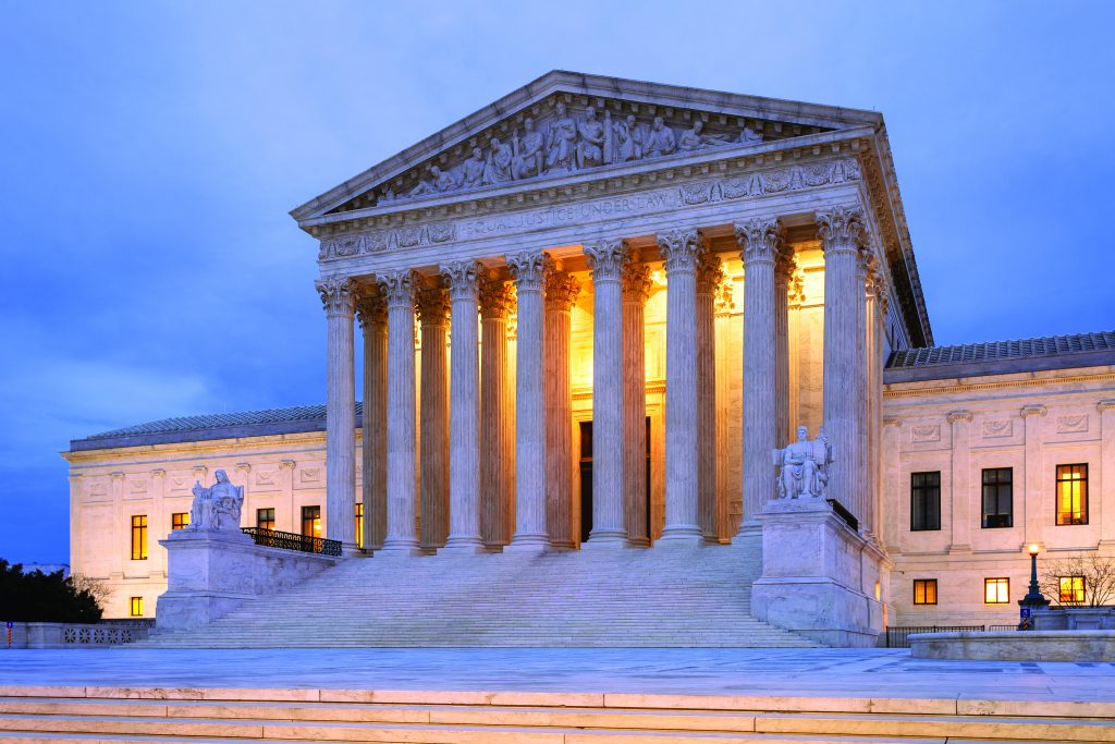 U.S. Supreme Court building at dusk with warm lighting, neoclassical design, grand staircase, tall columns, and statues prominently visible.