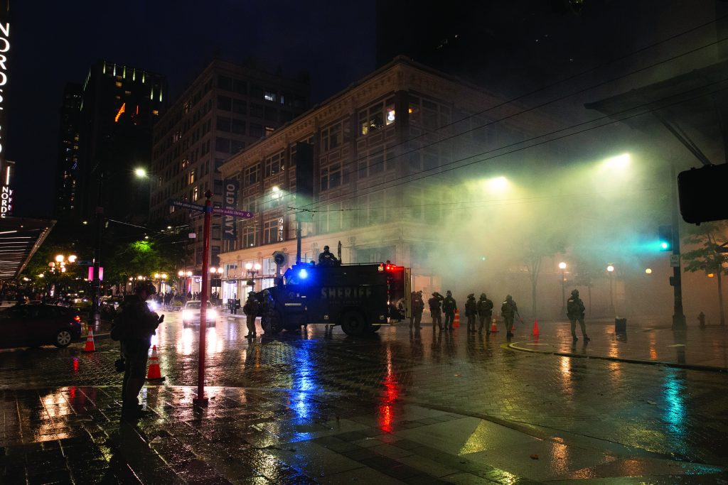 Night urban scene with officers, sheriff's armored vehicle flashing lights, smoke or tear gas, and wet streets reflecting city lights.