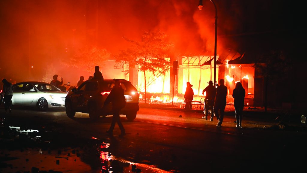 Nighttime blaze consumes building; bright orange flames light scene. Silhouetted people and two cars in foreground amid chaotic atmosphere.