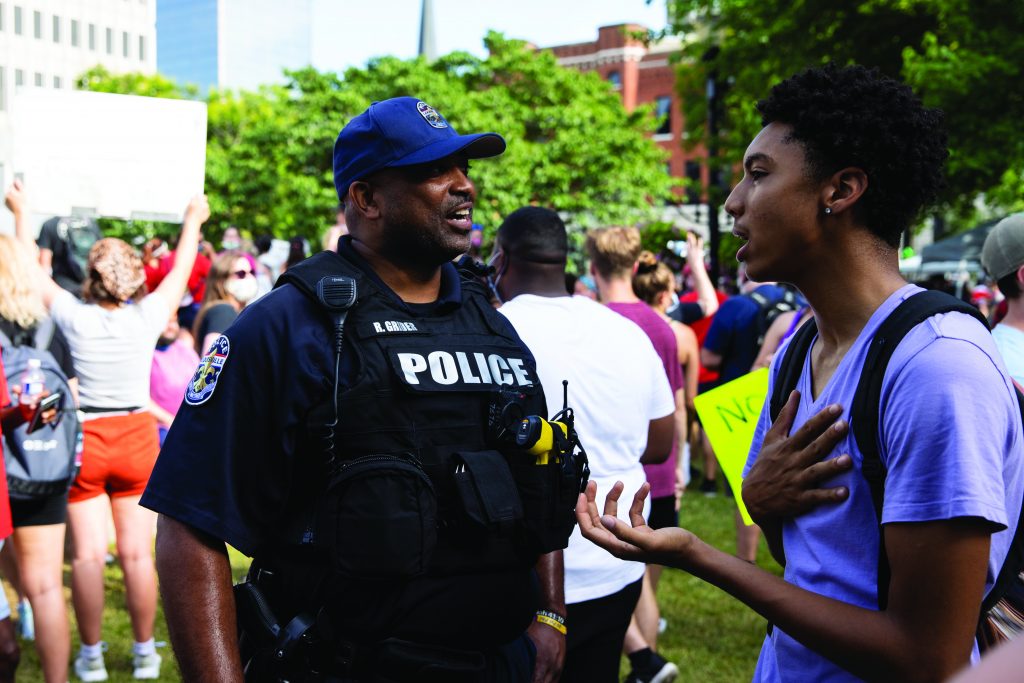 Police officer in tactical vest speaks with a person holding a hand to chest during outdoor protest; crowd with signs and city buildings in background.