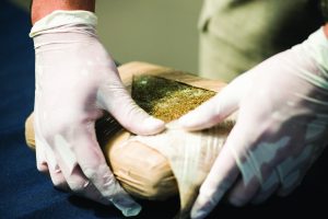 Gloved hands inspect plastic-wrapped rectangular package with greenish-brown substance inside, suggesting narcotics; blurred background for focus