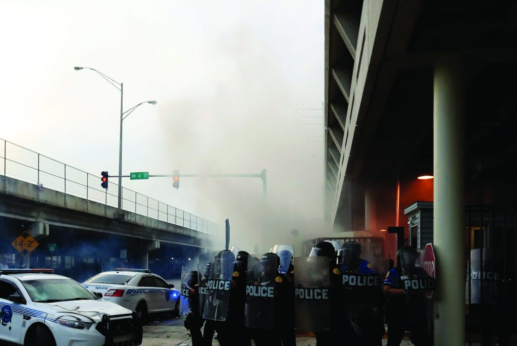 Officers in riot gear stand near graffitied police car; smoke fills air under highway overpass in tense urban setting.