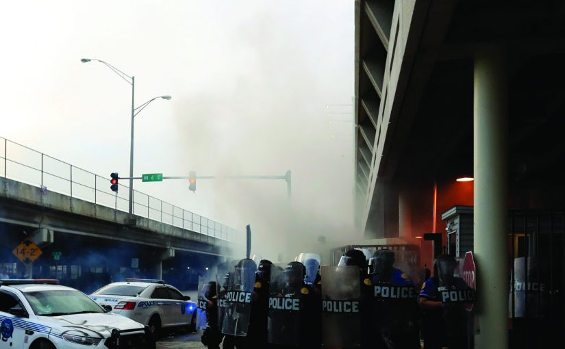 Officers in riot gear stand near graffitied police car; smoke fills air under highway overpass in tense urban setting.