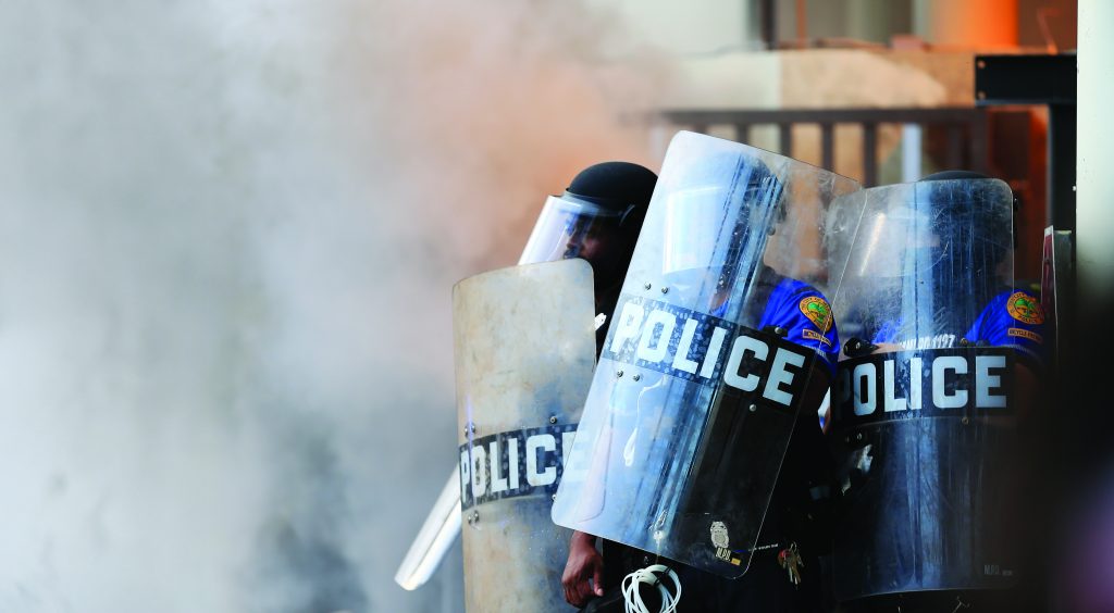 Three officers in riot gear hold clear 'POLICE' shields amid smoke-filled background, indicating tense or chaotic urban environment
