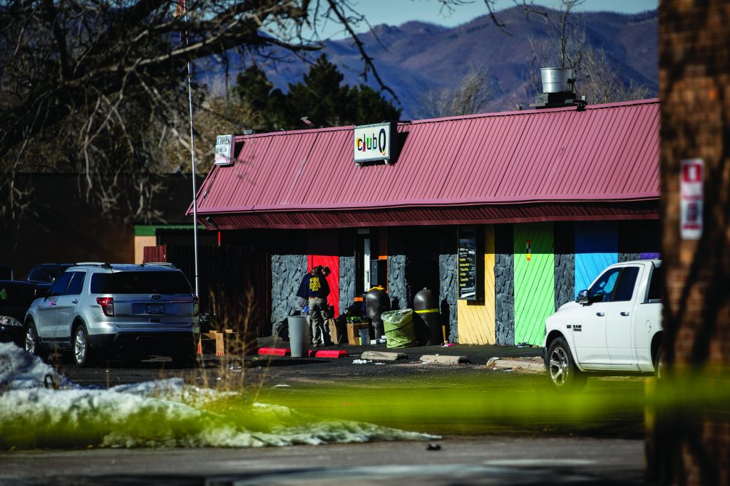Law enforcement vehicles and personnel stand outside a building with colorful boarded panels, crime‑scene tape in the foreground, and mountains in the distance.