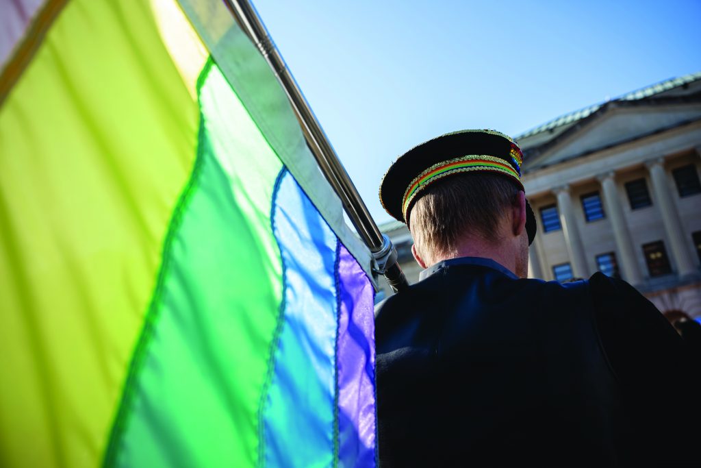 Uniformed officer stands near a large rainbow pride flag outside a government building with columns under clear blue sky, showing support for inclusivity.