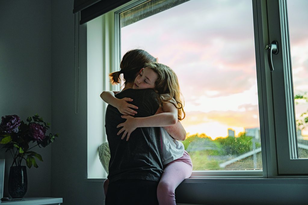 Child sitting on a person’s lap near a large window, arms wrapped around each other, with sunset view outside and flowers on a table in the foreground