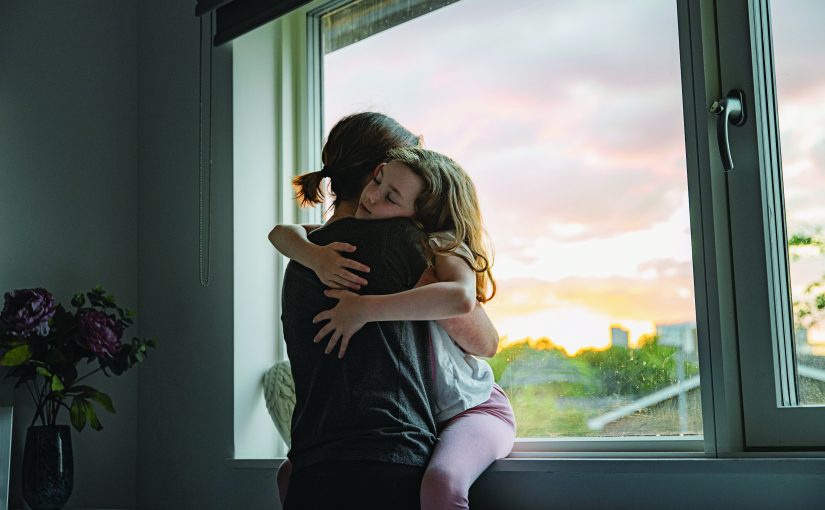 Child sitting on a person’s lap near a large window, arms wrapped around each other, with sunset view outside and flowers on a table in the foreground
