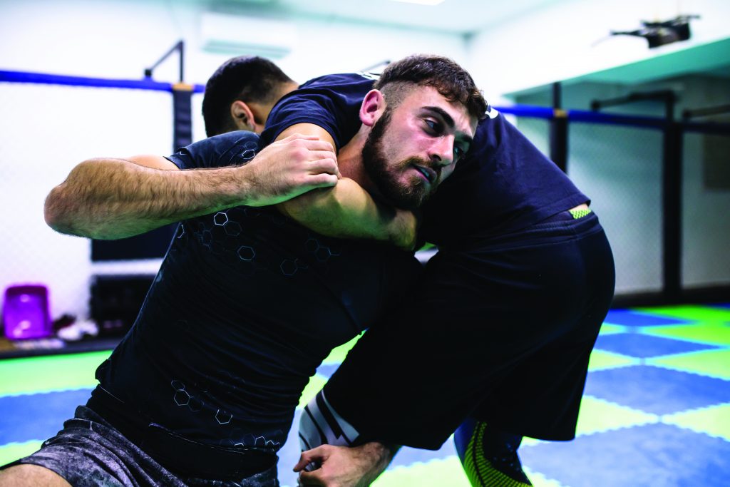 Two individuals grapple on blue-green checkered gym mat; one applies headlock during training, demonstrating physical control and defensive tactics.