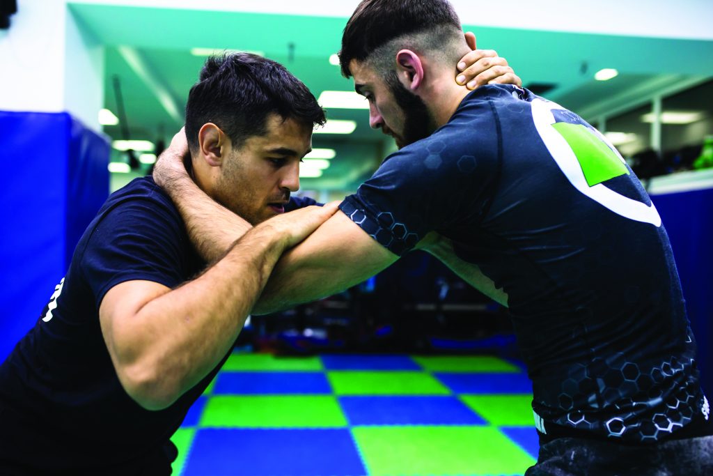 Two individuals in black athletic gear grapple on blue-green checkered mat in indoor facility; training stance emphasizes control and technique.