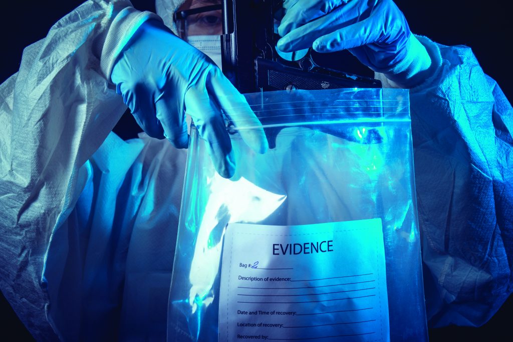 Forensic investigator in protective gear bags item in clear “EVIDENCE” pouch with labeled fields for ID, description, date, and location at scene.