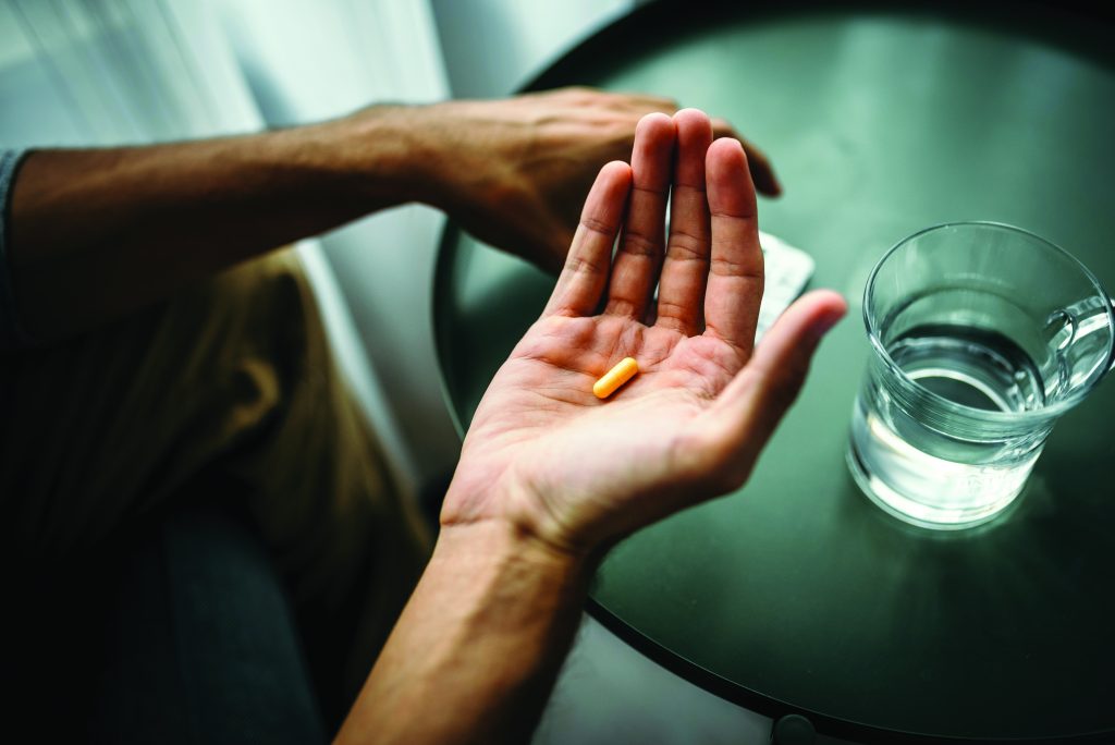 a close-up of a person's hand holding an orange capsule-shaped pill and a white round tablet. The hand is positioned over a dark green circular table. On the table, there is also a clear glass of water filled halfway. Another arm rests on the edge of the table, partially visible in the background. The faces are not visible in this image.
