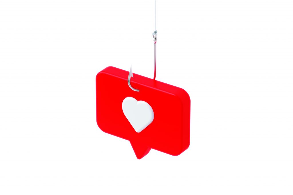 Red speech‑bubble icon with a white heart is pierced by a hanging fishhook, symbolizing social‑media phishing against a plain white background
