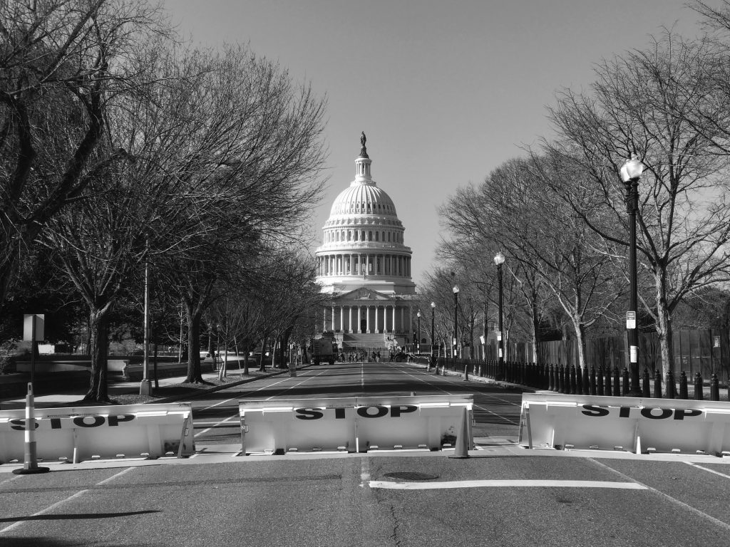 Black‑and‑white view of the U.S. Capitol centered at the end of an empty roadway blocked by ‘STOP’ barricades, lined with leafless trees, fencing, and street lamps