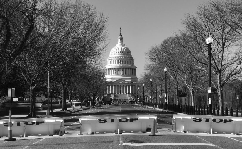 Black‑and‑white view of the U.S. Capitol centered at the end of an empty roadway blocked by ‘STOP’ barricades, lined with leafless trees, fencing, and street lamps