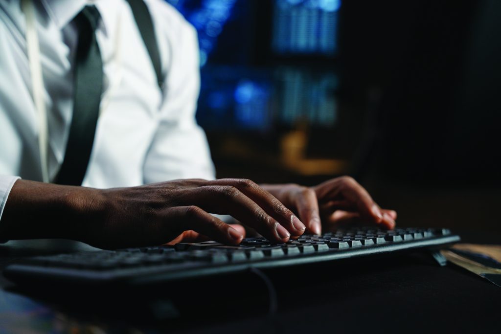 Person in white shirt and black tie typing on keyboard in dimly lit room with multiple computer screens displaying data and system interfaces