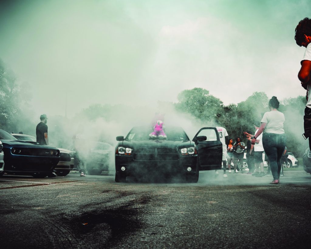 A dodge peeling out creating smoke as spectators watch. smoke takes up most of the picture along with people and the cars in the background.