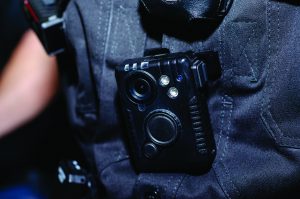 Close-up of body camera mounted on officer’s uniform; lens, buttons, and LED indicators visible against dark tactical fabric background.