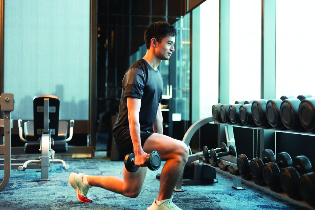 Person performing weighted lunge in gym, holding dumbbells in each hand near a rack of weights, with exercise equipment and large windows in background.