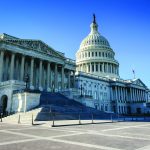 Wide exterior view of the U.S. Capitol building with white stone columns, broad steps, domed rotunda, empty plaza, and clear blue sky in daylight