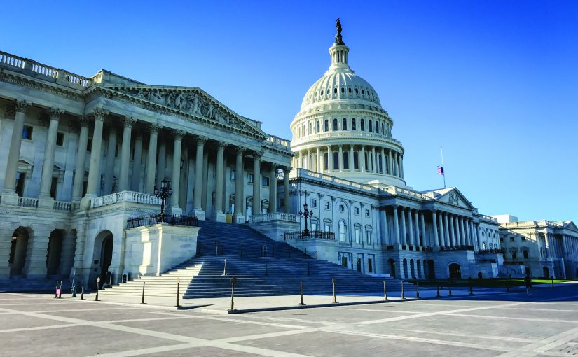 Wide exterior view of the U.S. Capitol building with white stone columns, broad steps, domed rotunda, empty plaza, and clear blue sky in daylight