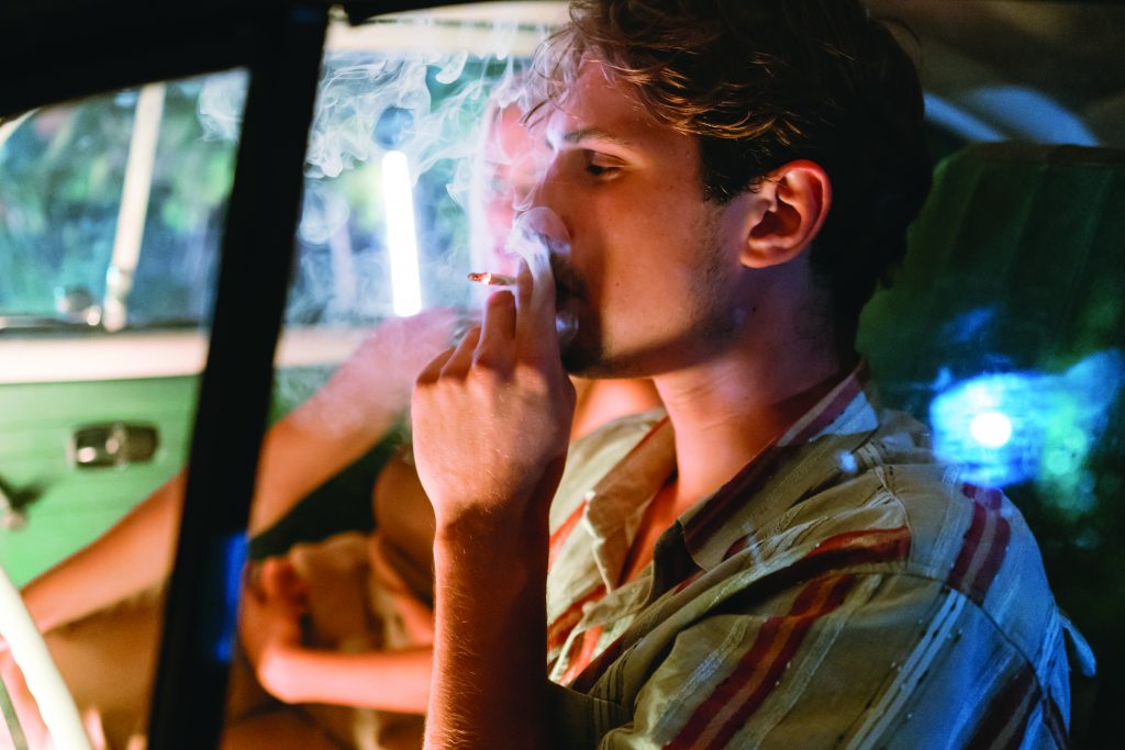 Individual in striped shirt smokes inside vehicle with window cracked; smoke surrounds hand and head; second person seated beside in front passenger seat.