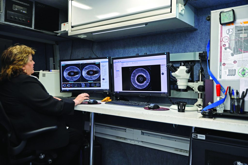 Technician at workstation reviewing circular forensic images on dual monitors beside a microscope, tools, notes, and equipment in a mobile lab setting
