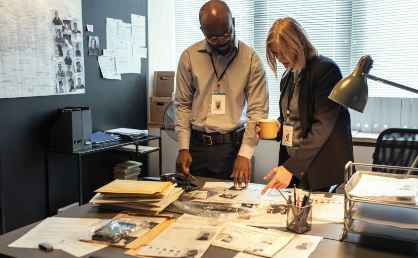 Two officers review documents and evidence on cluttered desk in office; lamp, case files, and wall photos suggest active investigation workspace.
