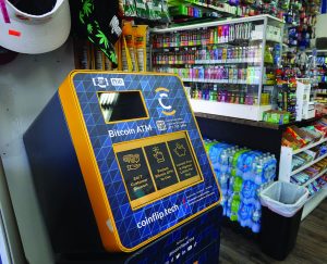 Bitcoin ATM kiosk with touchscreen, QR code, and instructions stands in a convenience store beside snacks, vape products, bottled water, and a checkout counter